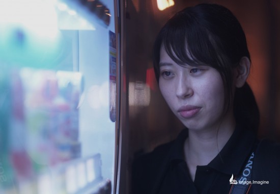 夜の街の中、明かりを放っている自動販売機を見つめている女性を撮影した写真。