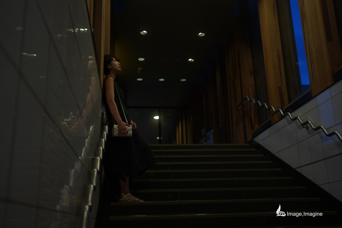 夜の街にて、地下へ続く階段で女性を撮影した写真。女性は全身黒いワンピースを着て、壁に寄り掛かりながら天井を見つめている。