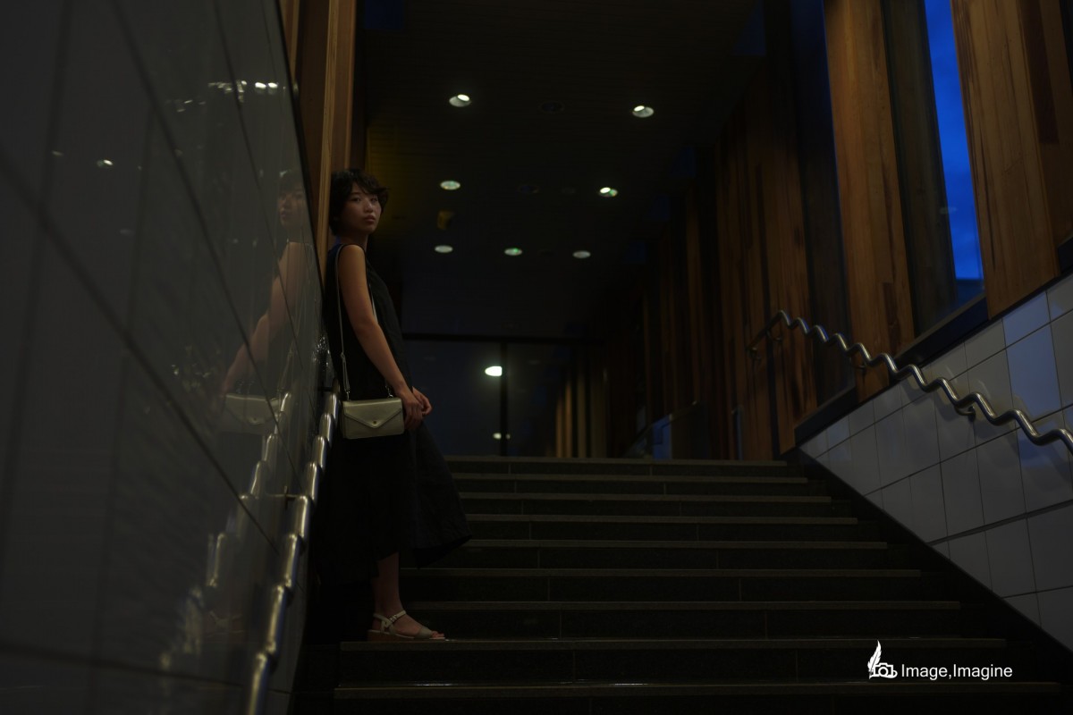 夜の街にて、地下へ続く階段で女性を撮影した写真。女性は全身黒いワンピースを着て、壁に寄り掛かりながらこちらを見つめている。