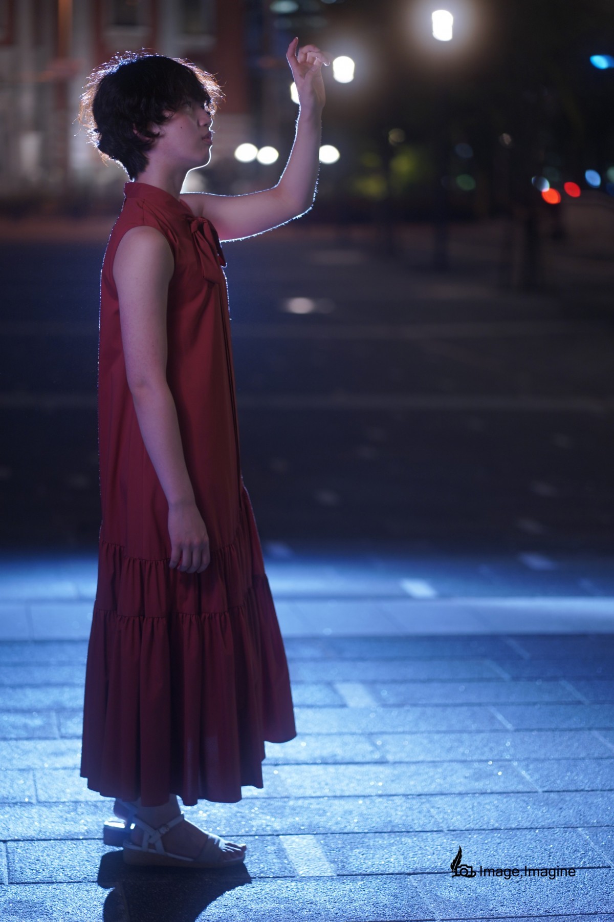 夜の街の中、赤いワンピースを着た女性を横から撮影した写真。女性は左手を空の方へ伸ばし、何かを掴もうとしている。