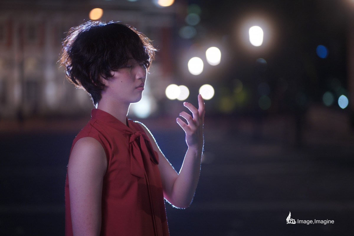 夜の街の中、赤いワンピースを着た女性を横から撮影した写真。女性は左の手のひらをじっと見つめている。
