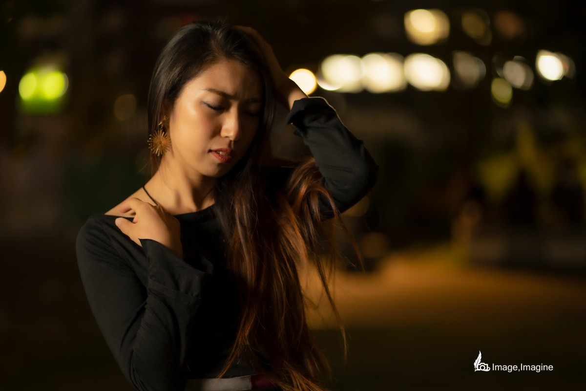 夜の街で女性を撮影した写真。女性は左手で髪をかき上げながら、少しうつむいている。