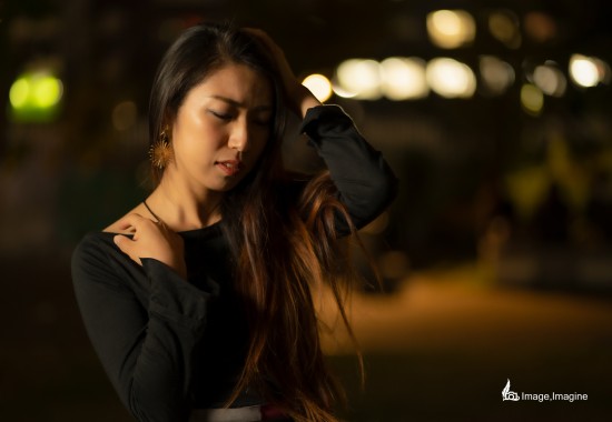 夜の街で女性を撮影した写真。女性は左手で髪をかき上げながら、少しうつむいている。