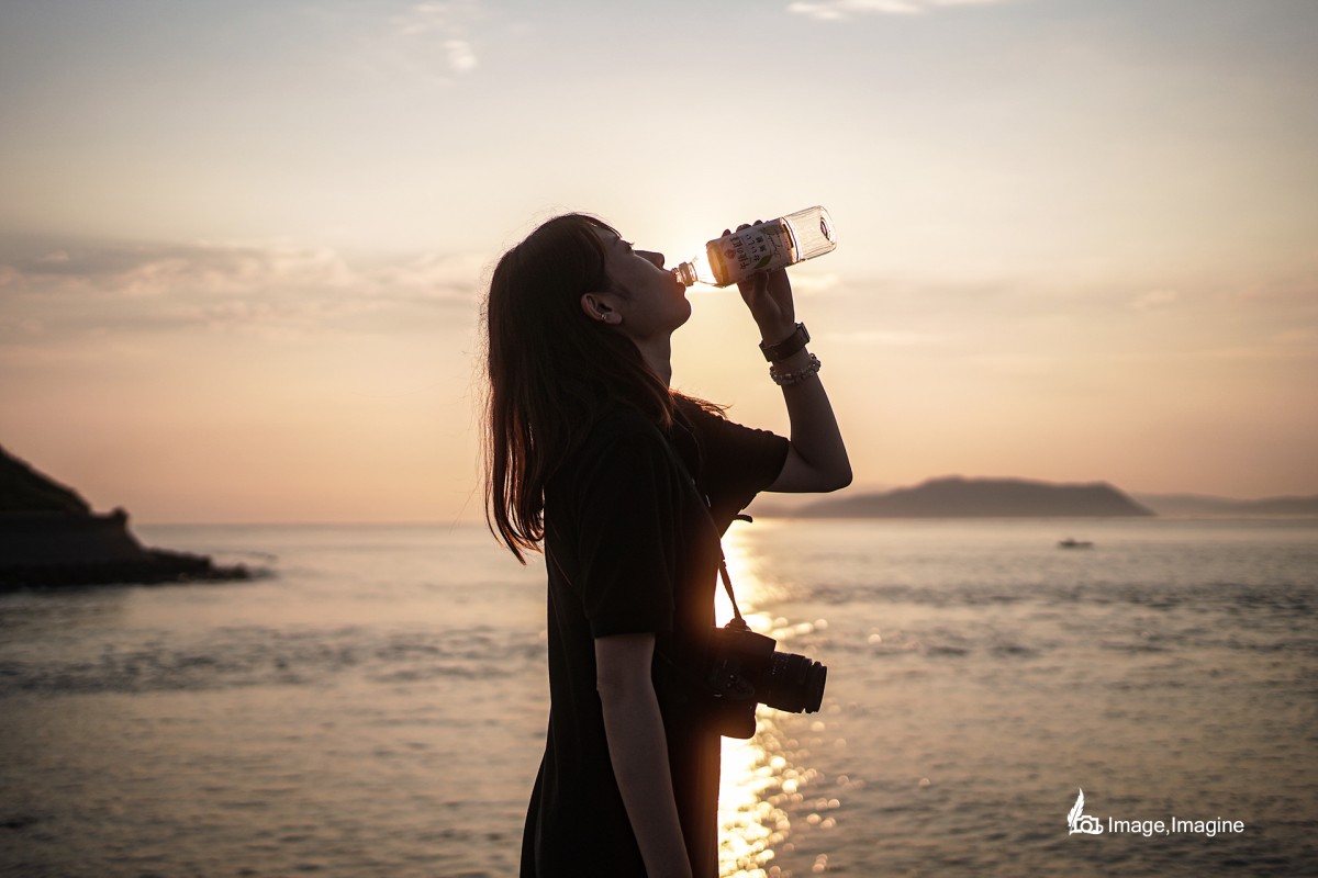 朝日と海を背景に女性を撮影した写真。女性はペットボトルに入った水を飲んでいる。
